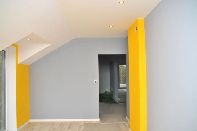 Wnętrze pokoju po malowaniu: szare ściany kontrastują z jaskrawożółtymi elementami, widoczne punkty oświetleniowe w suficie i fragment podłogi z jasnego drewna.