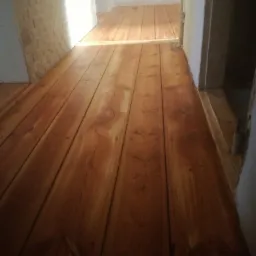 Lakierowana, drewniana podłoga z widocznymi słojami, prowadząca do jasnego pomieszczenia z ceglaną ścianą w tle.