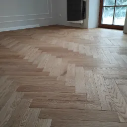 Nowa, jasna podłoga ułożona w jodełkę francuską, widoczny fragment ściany z listwami dekoracyjnymi, zasłonięty grzejnik i okno z widokiem na zimowy krajobraz.