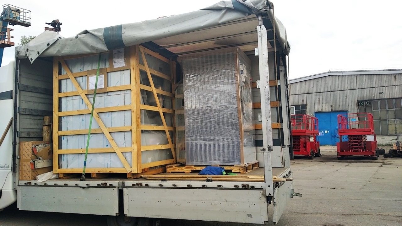 Załadunek dwóch dużych, drewnianych skrzyń i zapakowanego w folię urządzenia do wnętrza naczepy busa, z widocznymi niebieskimi pasami mocującymi i podnośnikami koszowymi w tle.