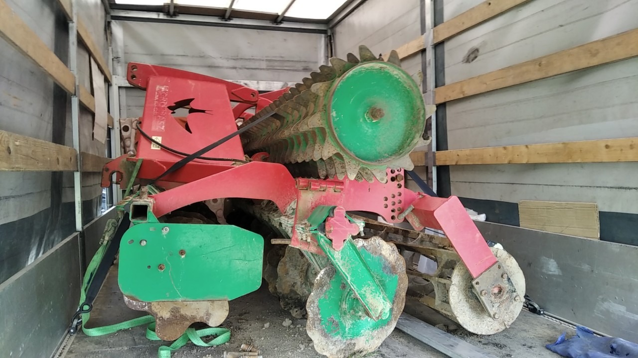 Czerwono-zielona brona talerzowa z logo jaskółki zabezpieczona pasami wewnątrz naczepy transportowej.