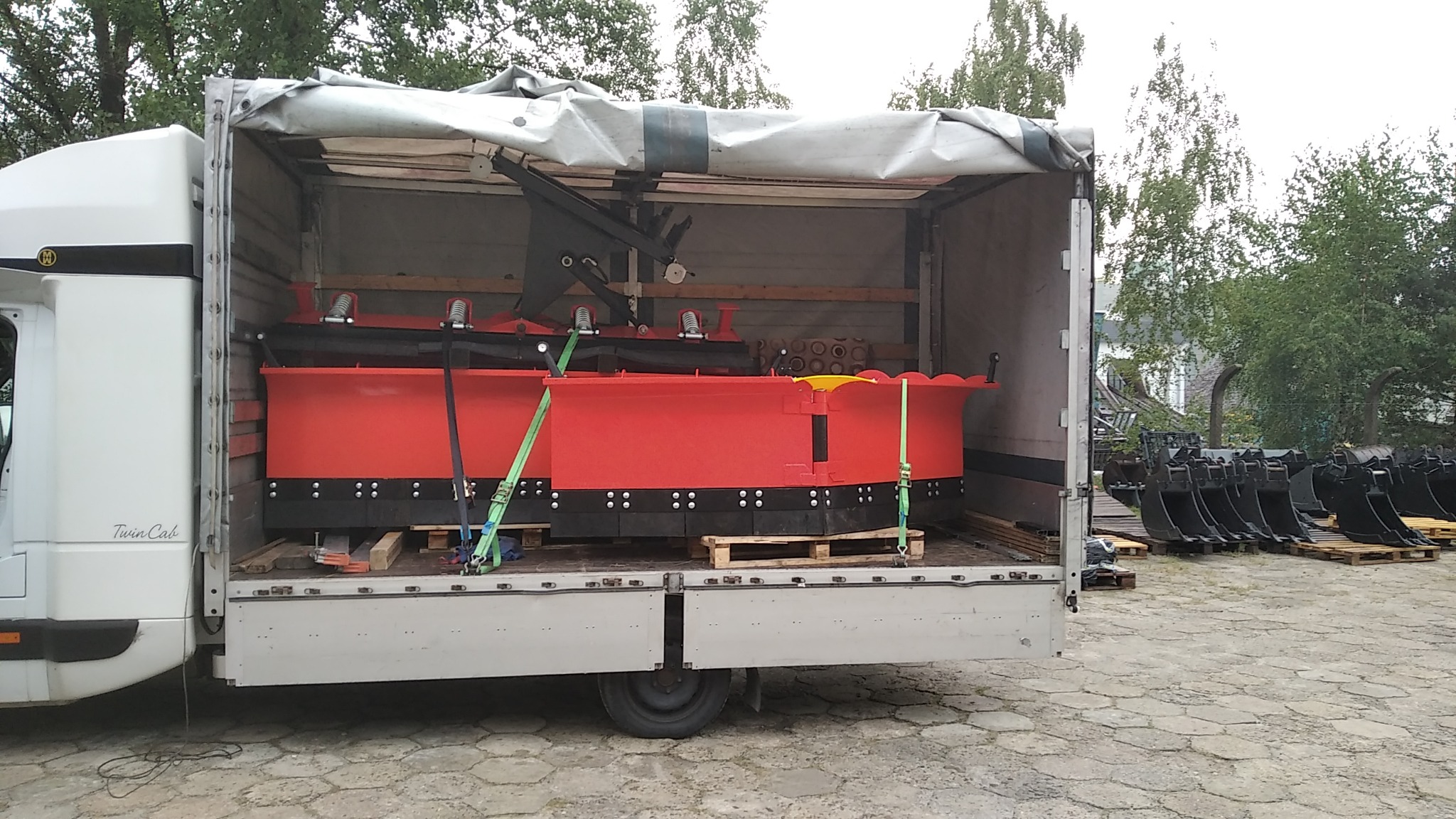 Czerwone pługi śnieżne zabezpieczone pasami wewnątrz otwartej naczepy transportowej, dodatkowe łyżki koparkowe na paletach w tle.