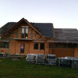 Niedokończony dom jednorodzinny z czerwonej cegły, częściowo pokryty dachówką, z oknami i drzwiami, na trawniku palety z materiałami budowlanymi.