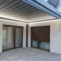 Nowoczesna pergola tarasowa z aluminiową konstrukcją i zintegrowanym oświetleniem LED, z widocznymi roletami zewnętrznymi i elementami wykończeniowymi w kolorze grafitowym.