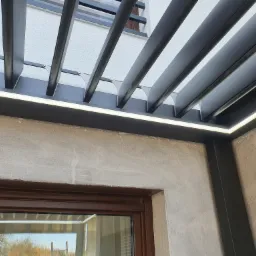 Nowoczesna pergola z metalowymi belkami i wbudowanym oświetleniem LED, widok z dołu na tle elewacji budynku i okna.