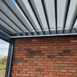 Nowoczesna pergola z szarymi, metalowymi lamelami i oświetleniem LED, przylegająca do ściany z czerwonej cegły z widocznym fragmentem krajobrazu w tle.