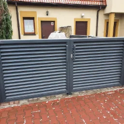 Szara, pozioma brama lamelowa z metalu, osadzona w kamiennych słupkach, przed budynkiem z beżową elewacją i czerwoną kostką brukową ze śniegiem.