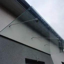 Szklany daszek nad wejściem do budynku, wsparty na metalowych wspornikach, widoczne krople deszczu na szkle.