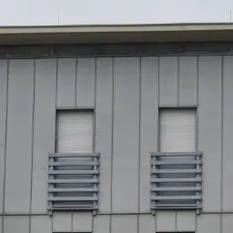 Fragment fasady budynku z szarej blachy z dwoma oknami z żaluzjami i metalowymi, poziomymi elementami dekoracyjnymi pod oknami.