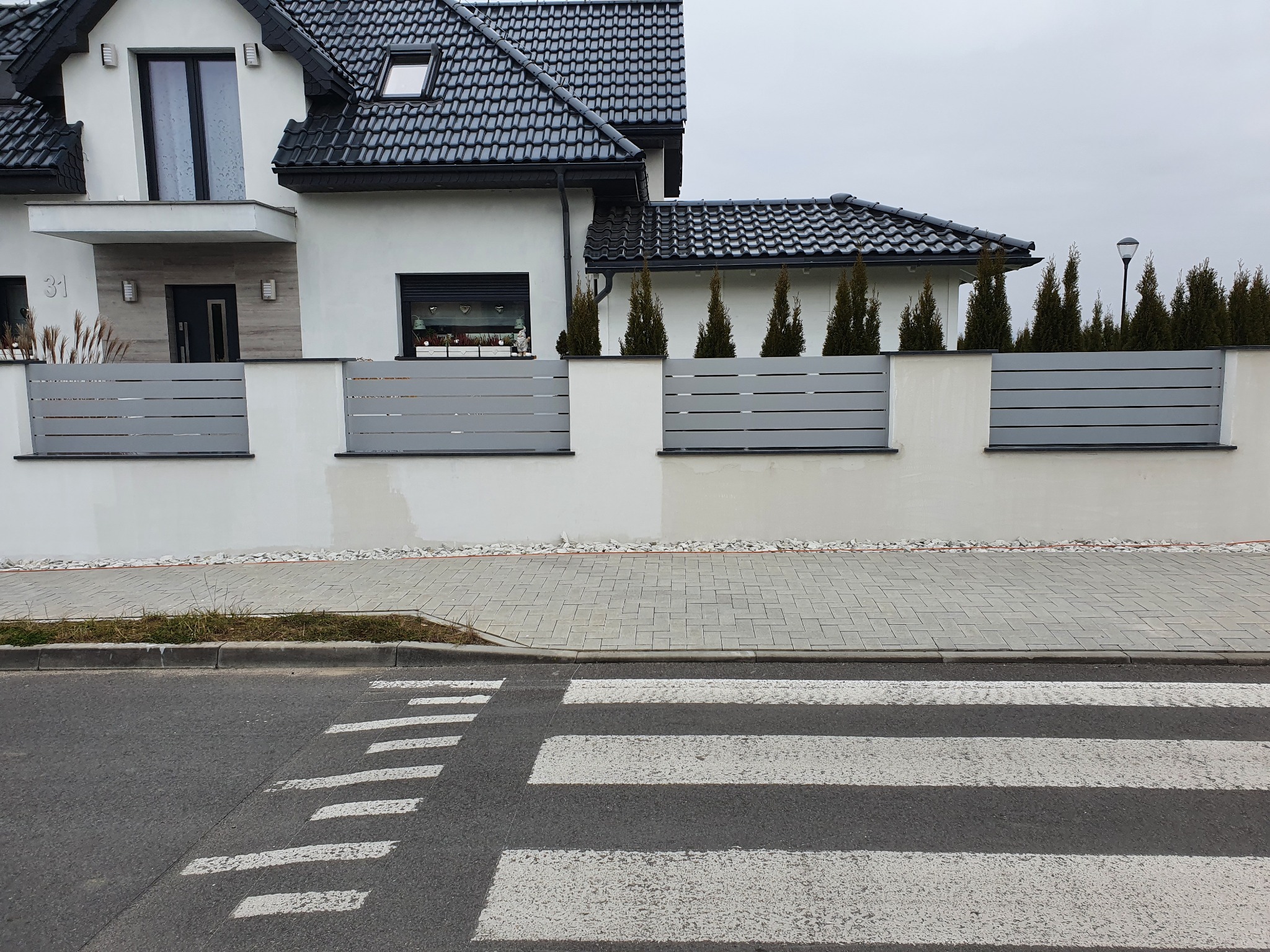 Nowoczesne ogrodzenie panelowe z poziomych szarych listew, oddzielające biały dom z czarnym dachem od ulicy z przejściem dla pieszych.