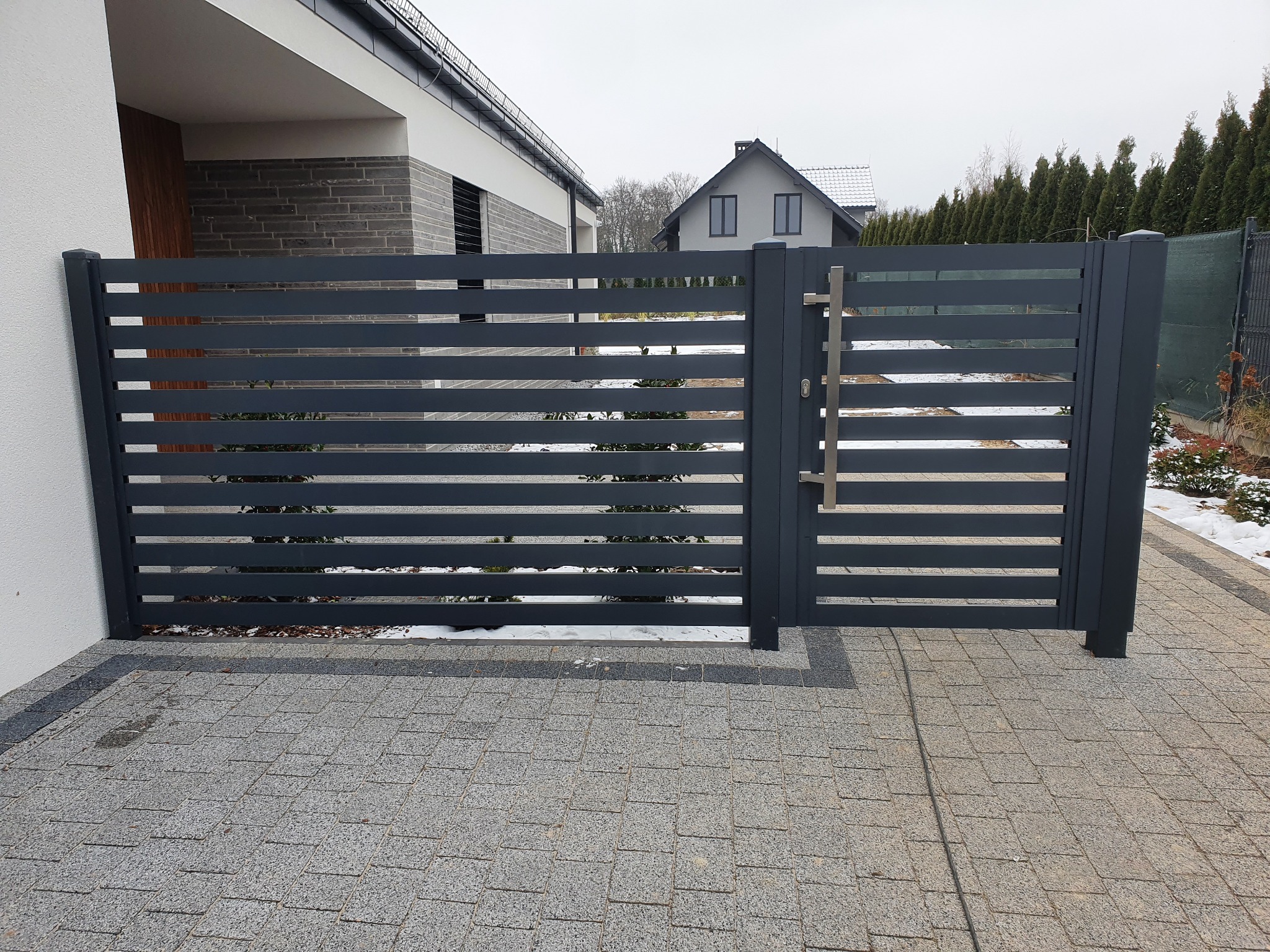 Nowoczesna, pozioma brama panelowa w kolorze grafitowym z metalowym uchwytem, zamontowana przy posesji z kostki brukowej, w tle dom i zimowy krajobraz.