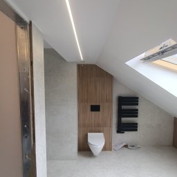 Usługi Remontowo Wykończeniowe Jarosław Rymarz - Wykończona łazienka na poddaszu ze skośnym sufitem, oświetleniem LED, drewnianą ścianą z toaletą i czarnym grzejnikiem drabinkowym, widok na otwarte okno dachowe, rama metalowa w lewym rogu.