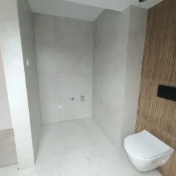 Nowoczesna łazienka z szarymi płytkami na ścianach i podłodze, drewniana ściana z zamontowaną miską WC, widoczne przyłącza wodne i elektryczne, sufit z otworem na oświetlenie.