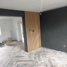 Montaż drewnianej ścianki działowej z listew na tle czarnej ściany w nowym mieszkaniu, widoczne kable elektryczne i ułożona podłoga w jodełkę.