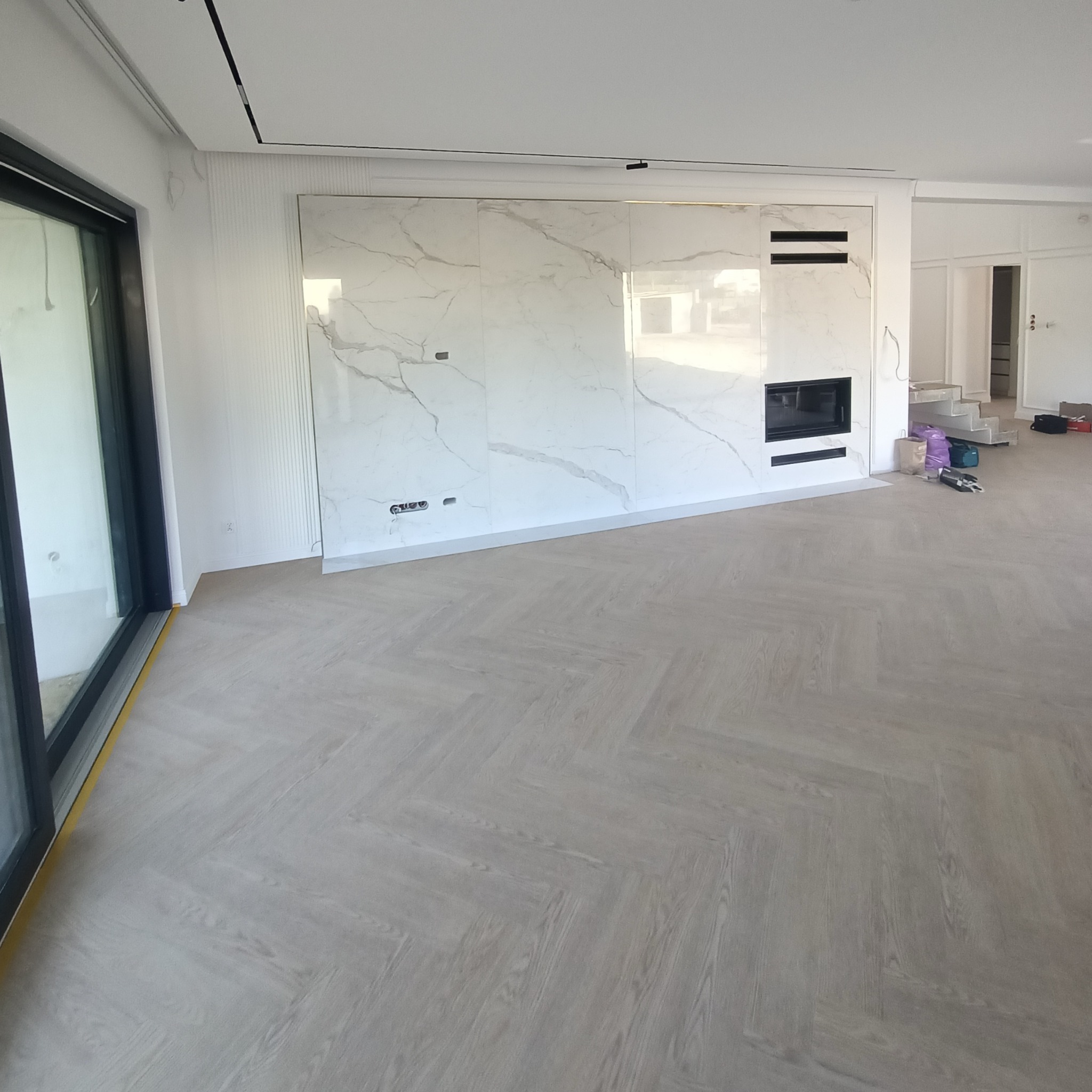 Salon po remoncie: parkiet w jodełkę, ściana z płyt imitujących marmur z wbudowanym kominkiem, duże okno przesuwne z czarną ramą, w tle schody.