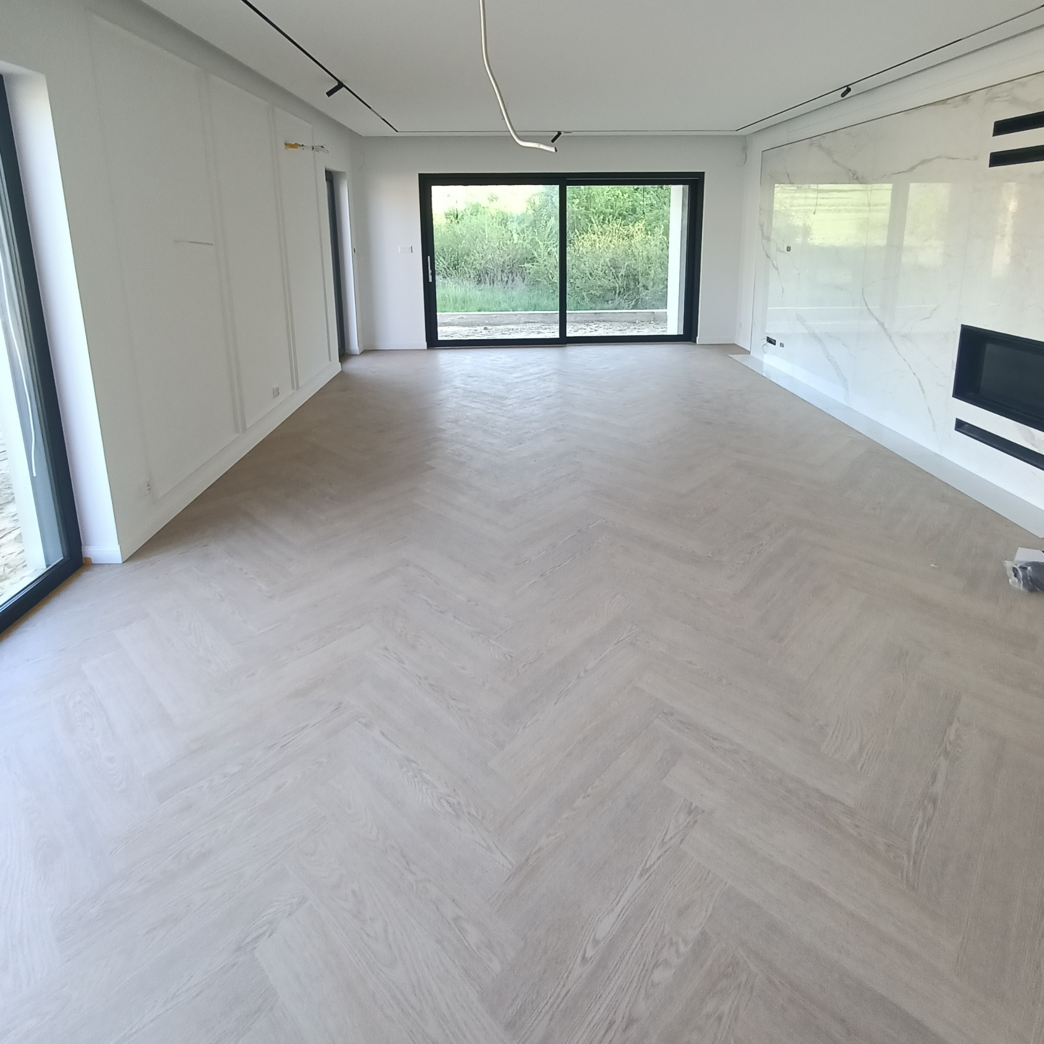 Jasny parkiet ułożony w jodełkę w przestronnym, nowoczesnym salonie z dużymi oknami przesuwnymi i marmurową ścianą z kominkiem.