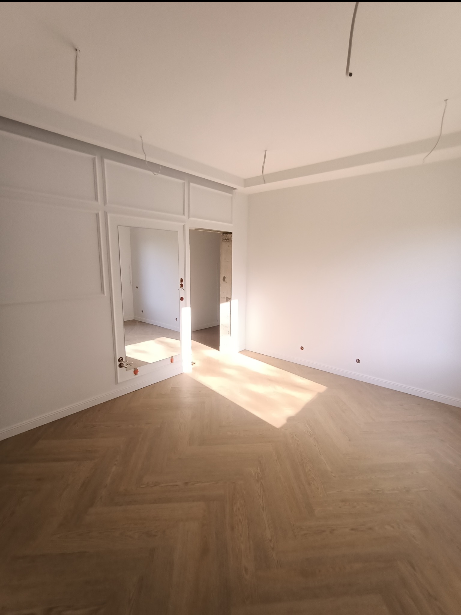 Pusty pokój po remoncie z parkietem ułożonym w jodełkę, białe ściany z dekoracyjnymi listwami i lustrem wbudowanym w ścianę, widoczne wystające kable z sufitu i przygotowane otwory na gniazdka.