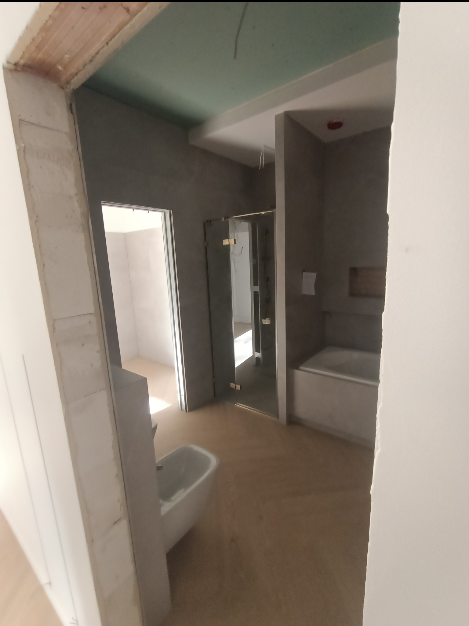 Łazienka w trakcie remontu: surowy beton na ścianach, widoczna wanna, kabina prysznicowa z przezroczystymi drzwiami, umywalka i podłoga z jasnego drewna. Ościeżnica drzwi w stanie surowym.