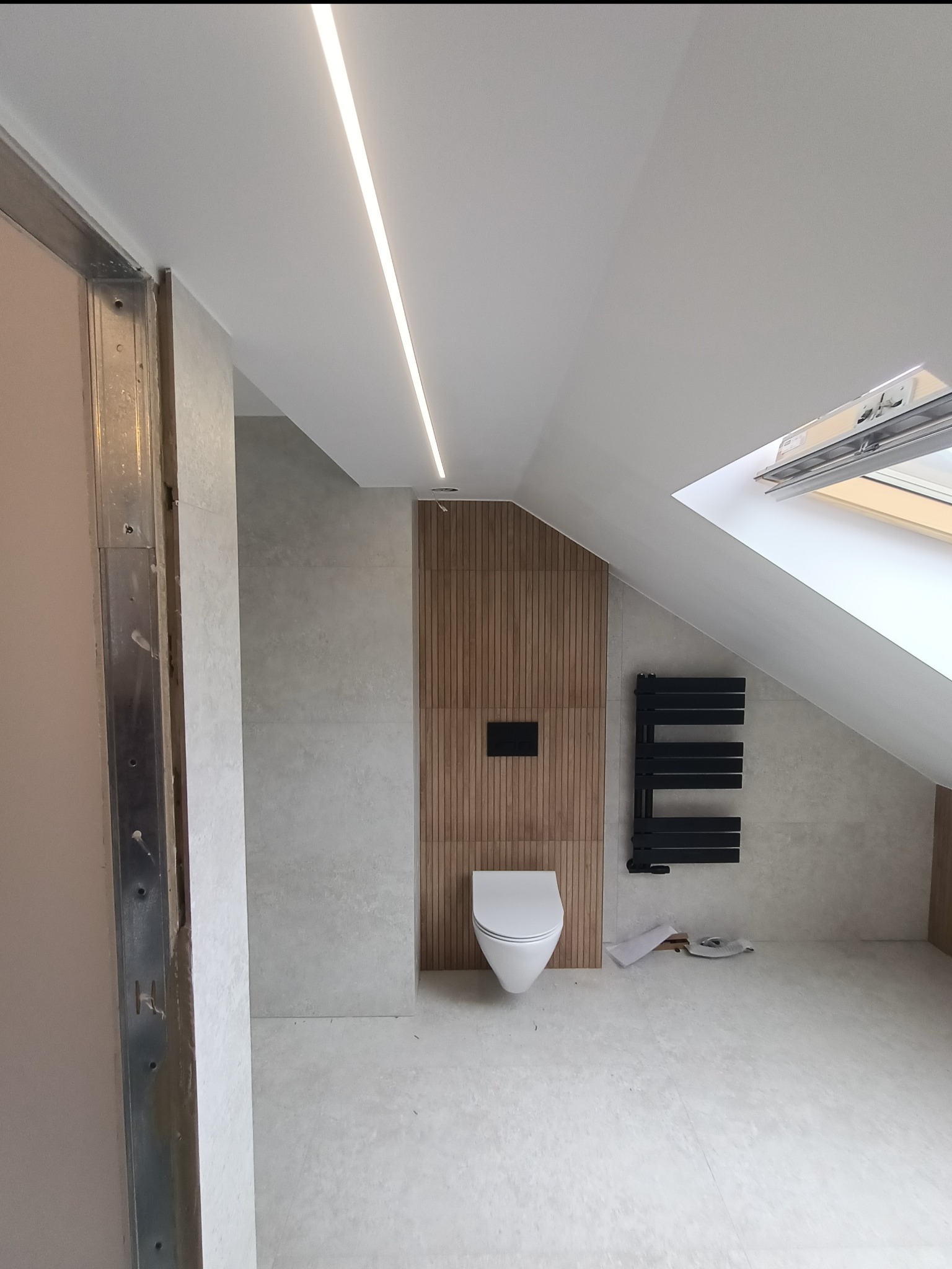 Wykończona łazienka na poddaszu ze skośnym sufitem, oświetleniem LED, drewnianą ścianą z toaletą i czarnym grzejnikiem drabinkowym, widok na otwarte okno dachowe, rama metalowa w lewym rogu.