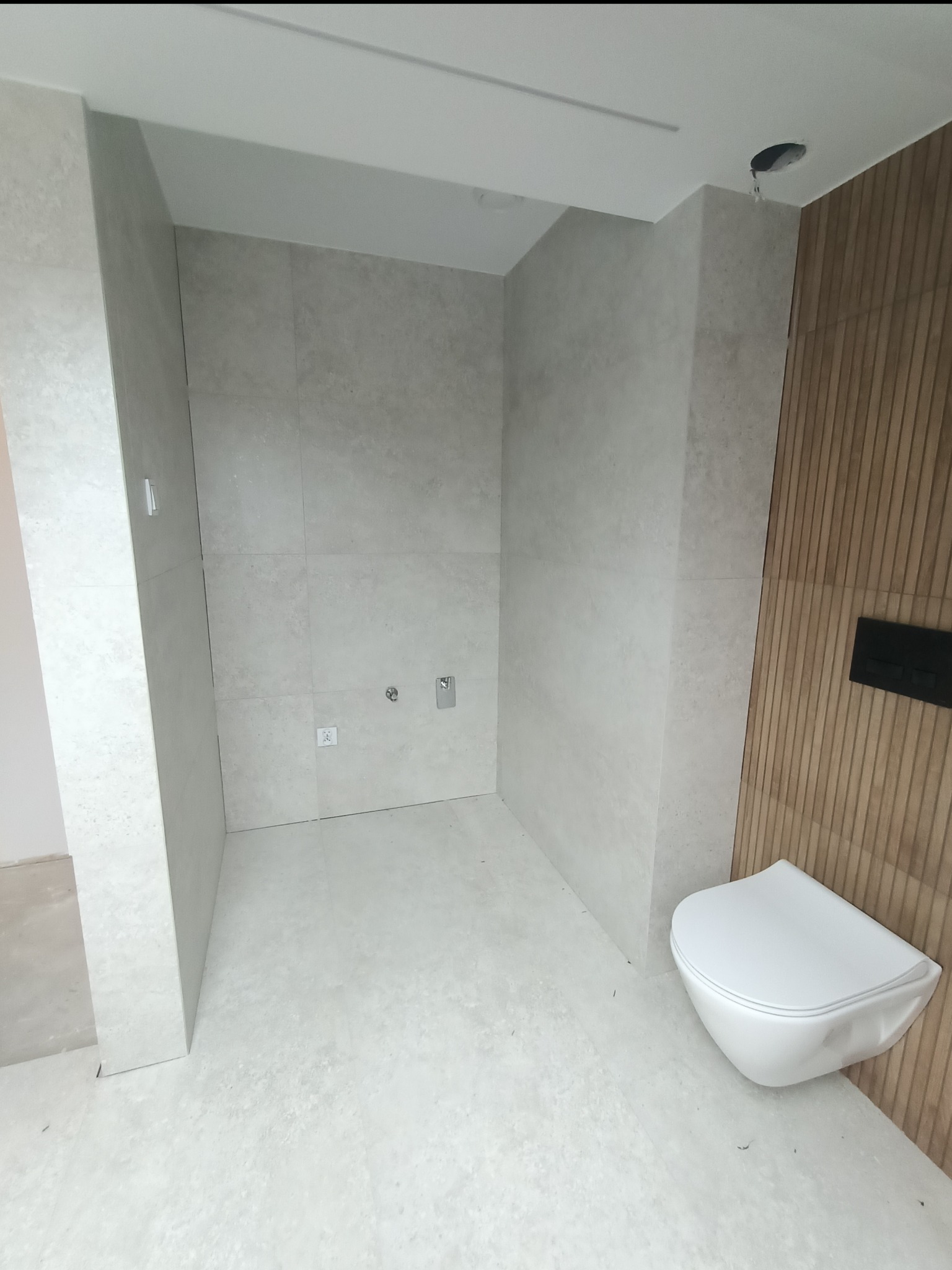 Nowoczesna łazienka z szarymi płytkami na ścianach i podłodze, drewniana ściana z zamontowaną miską WC, widoczne przyłącza wodne i elektryczne, sufit z otworem na oświetlenie.