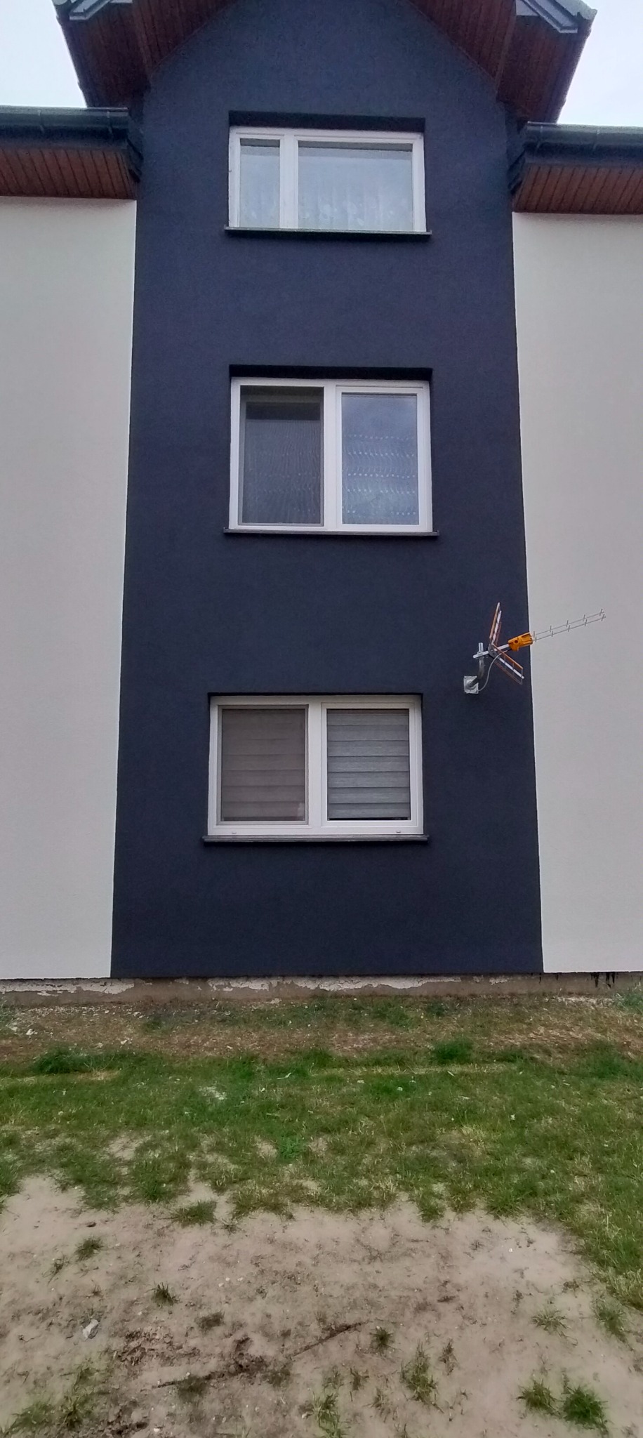 Fragment dwukolorowej elewacji budynku z trzema oknami, na ciemnej części elewacji widoczna antena satelitarna