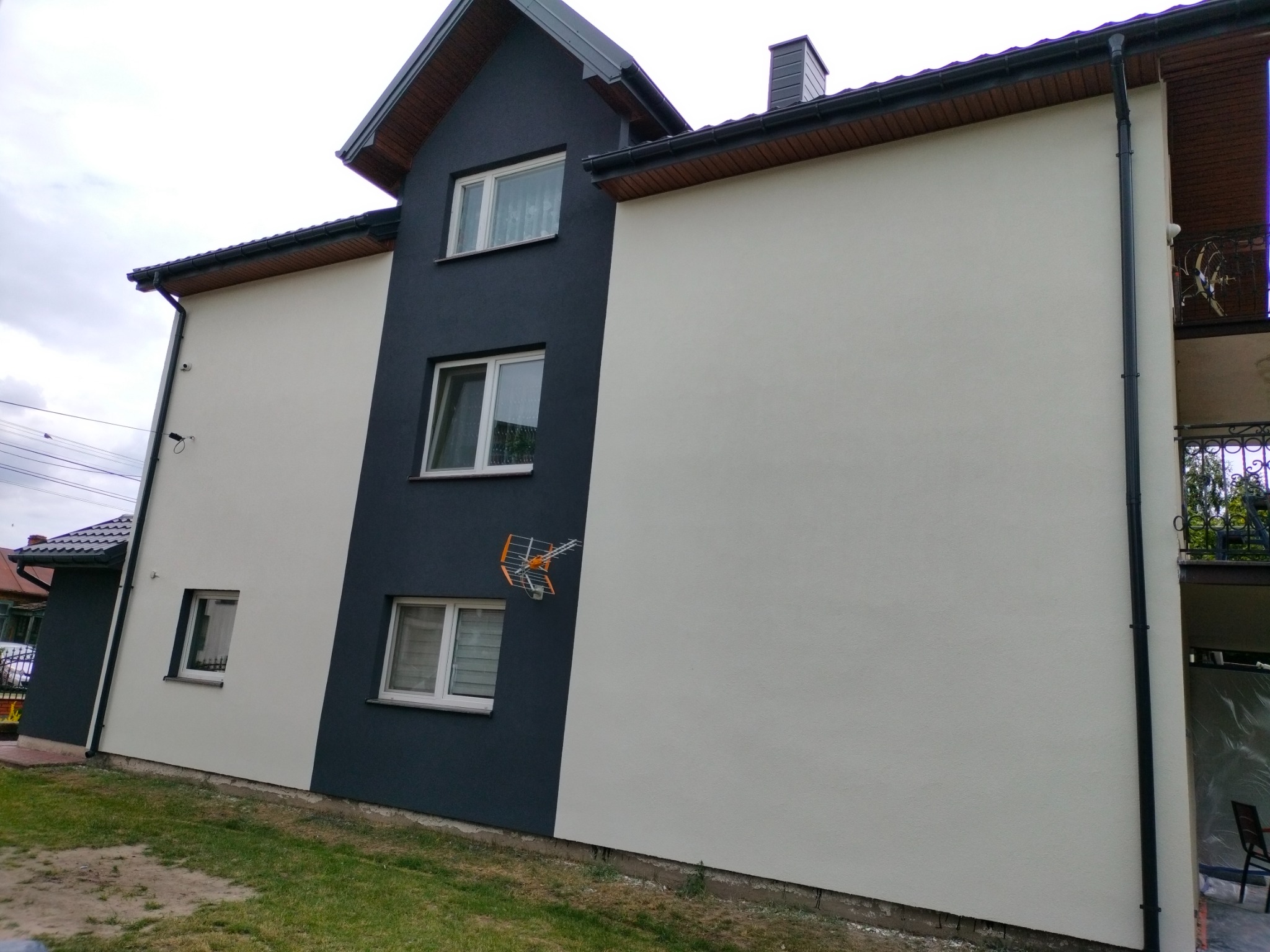 Dwukolorowa elewacja domu jednorodzinnego w Pniewie, połączenie jasnej szarości i grafitu, widoczna antena telewizyjna na ścianie.