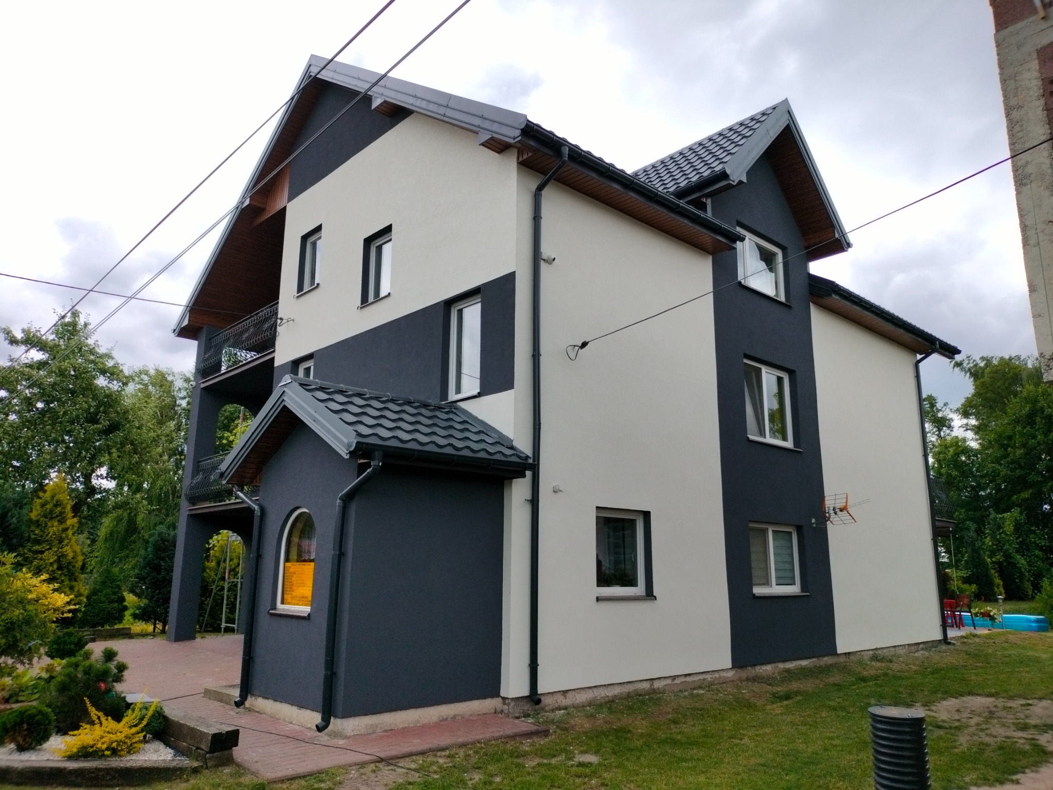 Dwukondygnacyjny dom z elewacją w kolorze białym i grafitowym, z balkonem i przyległą dobudówką, widok z zewnątrz w pochmurny dzień.