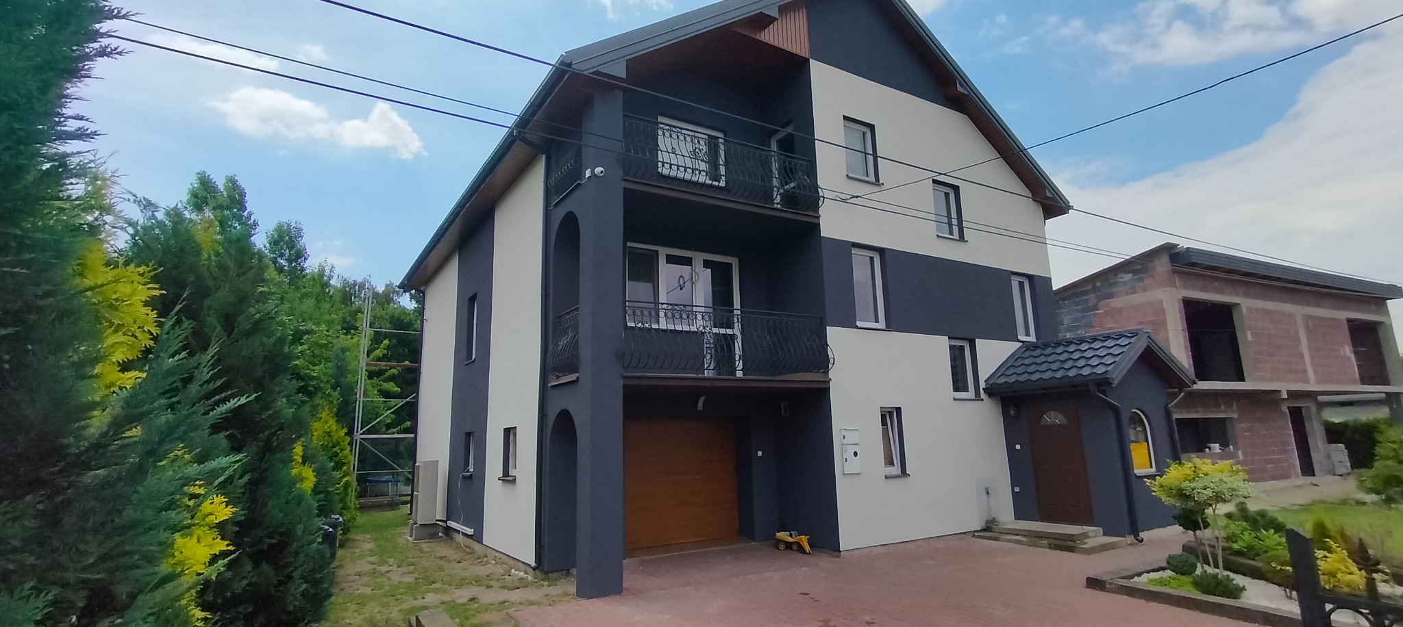 Dwukondygnacyjny dom z elewacją w odcieniach szarości i bieli, z balkonami z kutą balustradą, widok z zewnątrz na tle zieleni i częściowo wykończonego budynku.