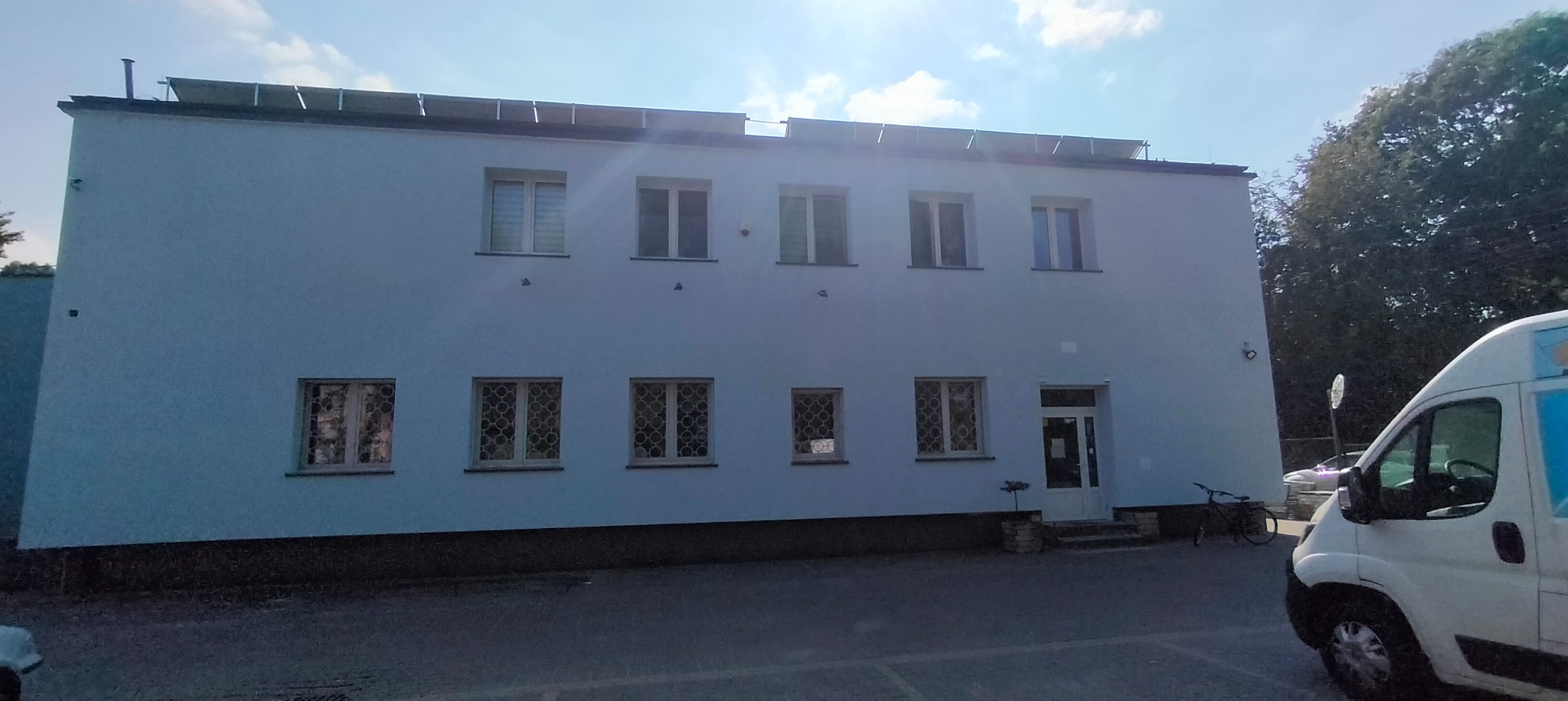 Dwukondygnacyjny budynek z jasną elewacją i panelami słonecznymi na dachu, widoczny z poziomu parkingu, z fragmentem białego samochodu dostawczego w prawym dolnym rogu.