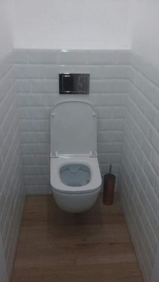 Wąska toaleta z białymi, cegiełkowymi płytkami na ścianach i drewnopodobną podłogą, wisząca muszla klozetowa z otwartą klapą, chromowany przycisk spłuczki i szczotka do WC w rogu.