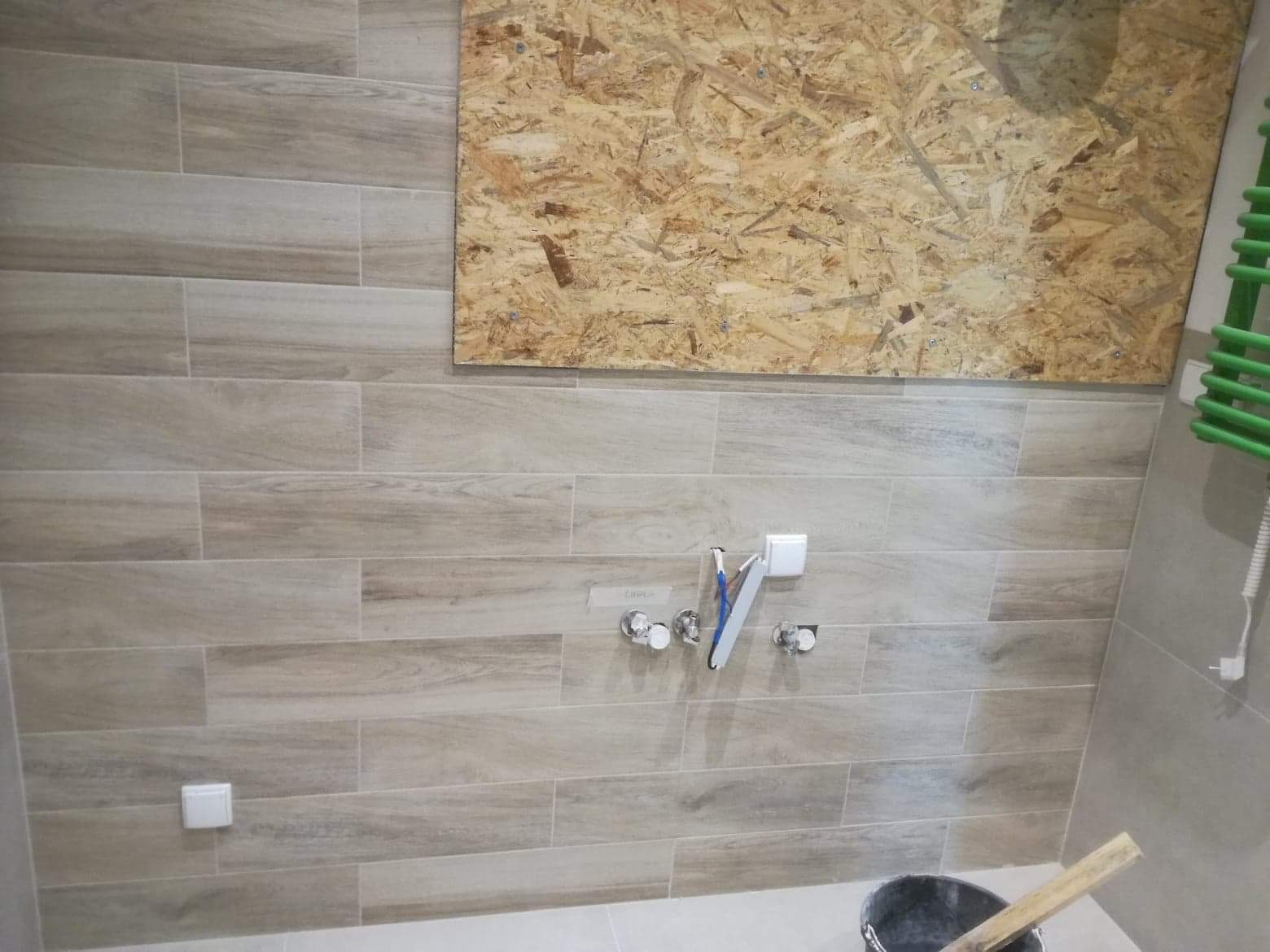 Ściana w trakcie układania płytek imitujących drewno, widoczne przyłącza wodne i elektryczne, zabezpieczona płyta OSB na górze ściany oraz wiadro z zaprawą i zielony grzejnik drabinkowy.