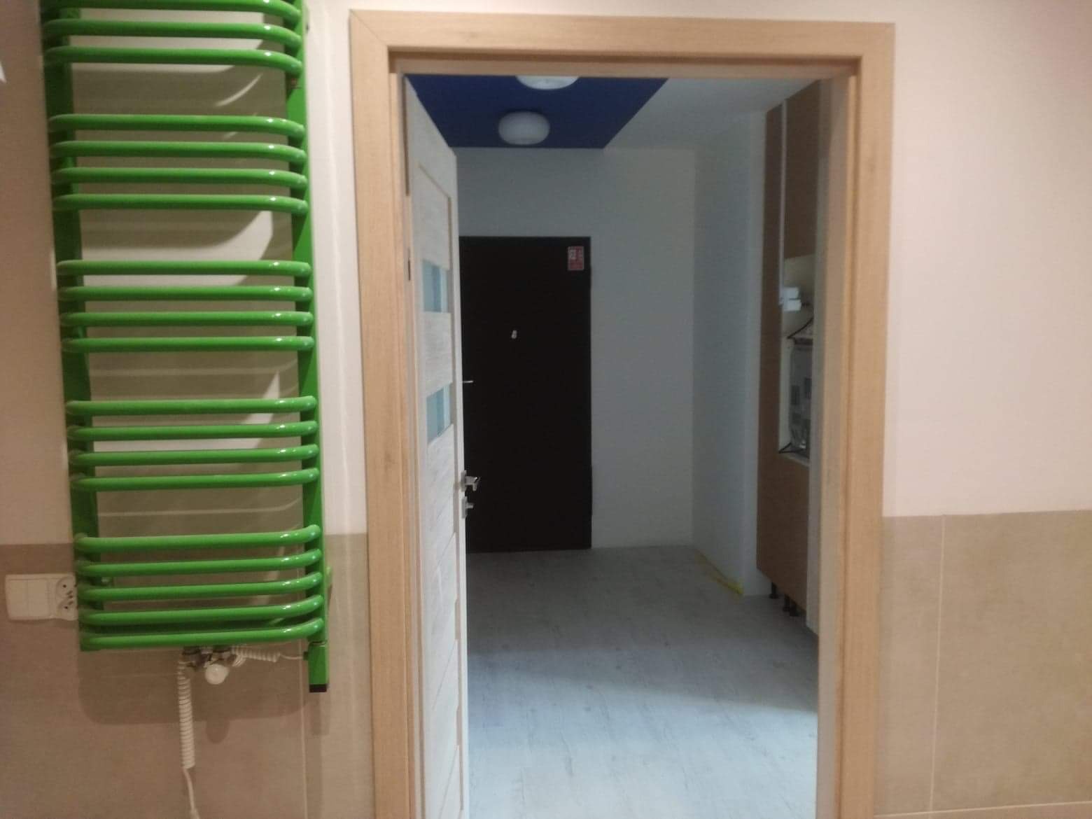 Zielony, drabinkowy grzejnik łazienkowy zamontowany na ścianie z beżowymi płytkami, widoczny fragment otwartych drzwi z jasną ościeżnicą i ciemnymi drzwiami wejściowymi w tle.