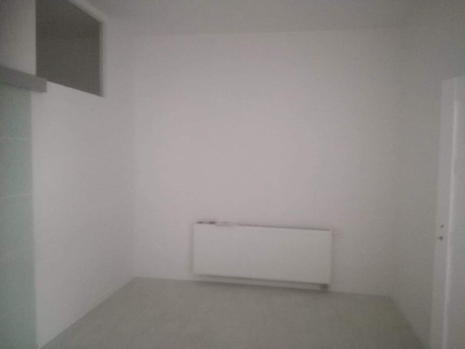 Pusty, pomalowany na biało pokój z grzejnikiem panelowym i drzwiami po prawej stronie, przygotowany do dalszych prac wykończeniowych.