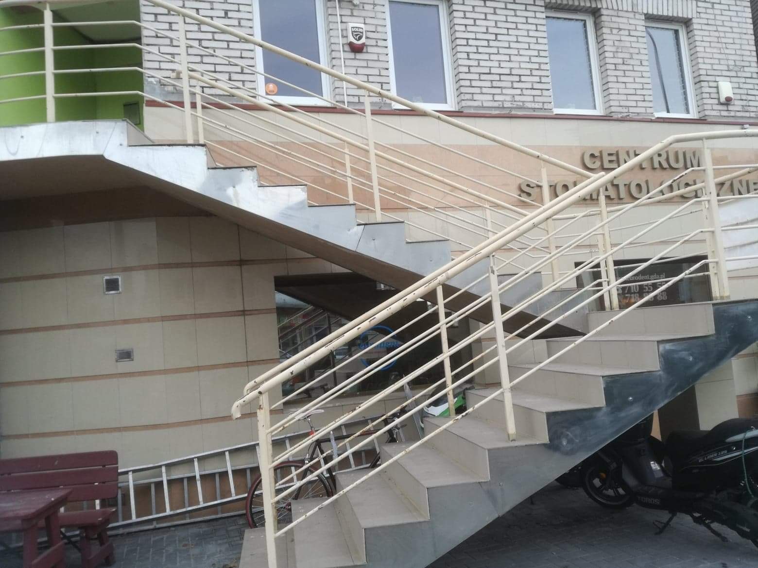 Zewnętrzne schody z kremowymi barierkami prowadzące do Centrum Stomatologicznego, obok zaparkowany skuter i rower, w tle drabina oparta o ścianę.