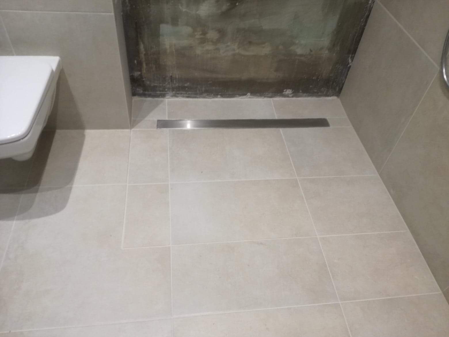 Ukończona łazienka z beżowymi płytkami podłogowymi i ściennymi, widoczny fragment podwieszanej toalety oraz liniowy odpływ pod prysznicem, ściana prysznica w trakcie wykończenia.