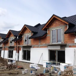 Budynek w trakcie ocieplania elewacji, widoczne płyty styropianowe, cegły, czarne dachy, okna i materiały budowlane porozrzucane na ziemi.