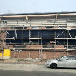 LEMBERG Group - Budynek z cegły w trakcie ocieplania, osłonięty rusztowaniem, widoczne okna zabezpieczone folią, żółta skrzynka elektryczna i zaparkowany srebrny samochód.