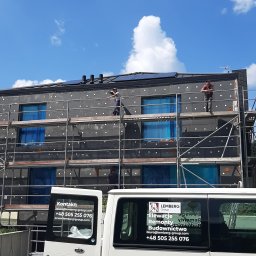 LEMBERG Group - Ocieplanie elewacji budynku wielorodzinnego z użyciem rusztowania, widoczne płyty styropianowe z kołkami, okna zabezpieczone folią, na dachu panele słoneczne, pracownicy w trakcie pracy,...