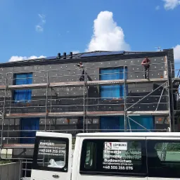 Ocieplanie elewacji budynku wielorodzinnego z użyciem rusztowania, widoczne płyty styropianowe z kołkami, okna zabezpieczone folią, na dachu panele słoneczne, pracownicy w trakcie pracy,...