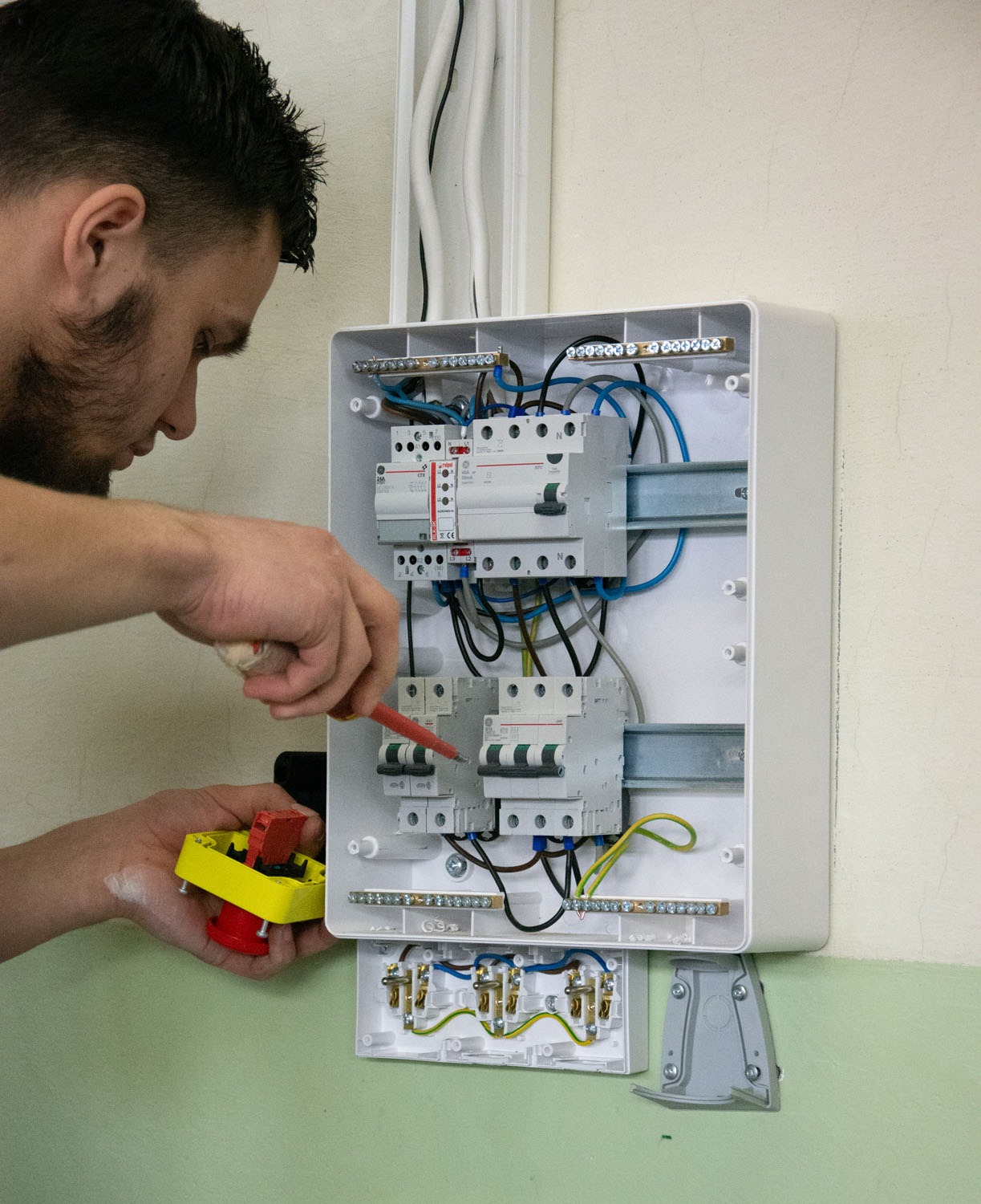 Elektryk z miernikiem napięcia i wyłącznikiem w rękach, pracuje przy otwartej skrzynce z bezpiecznikami i przewodami wewnątrz budynku.