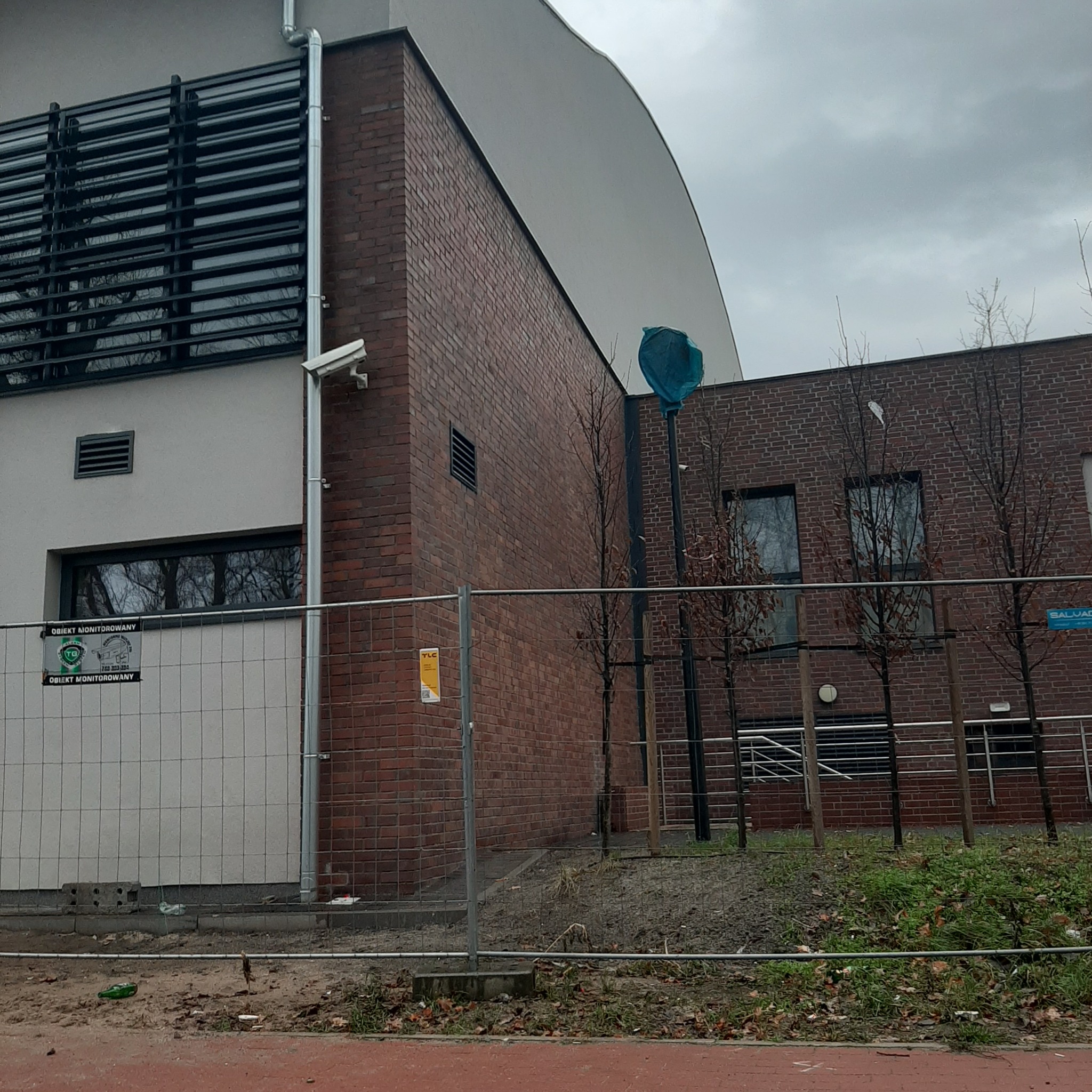 Budynek z cegły i białego tynku z widocznymi elementami instalacji elektrycznych: kamera monitoringu i rura spustowa. Całość otoczona tymczasowym ogrodzeniem budowlanym.