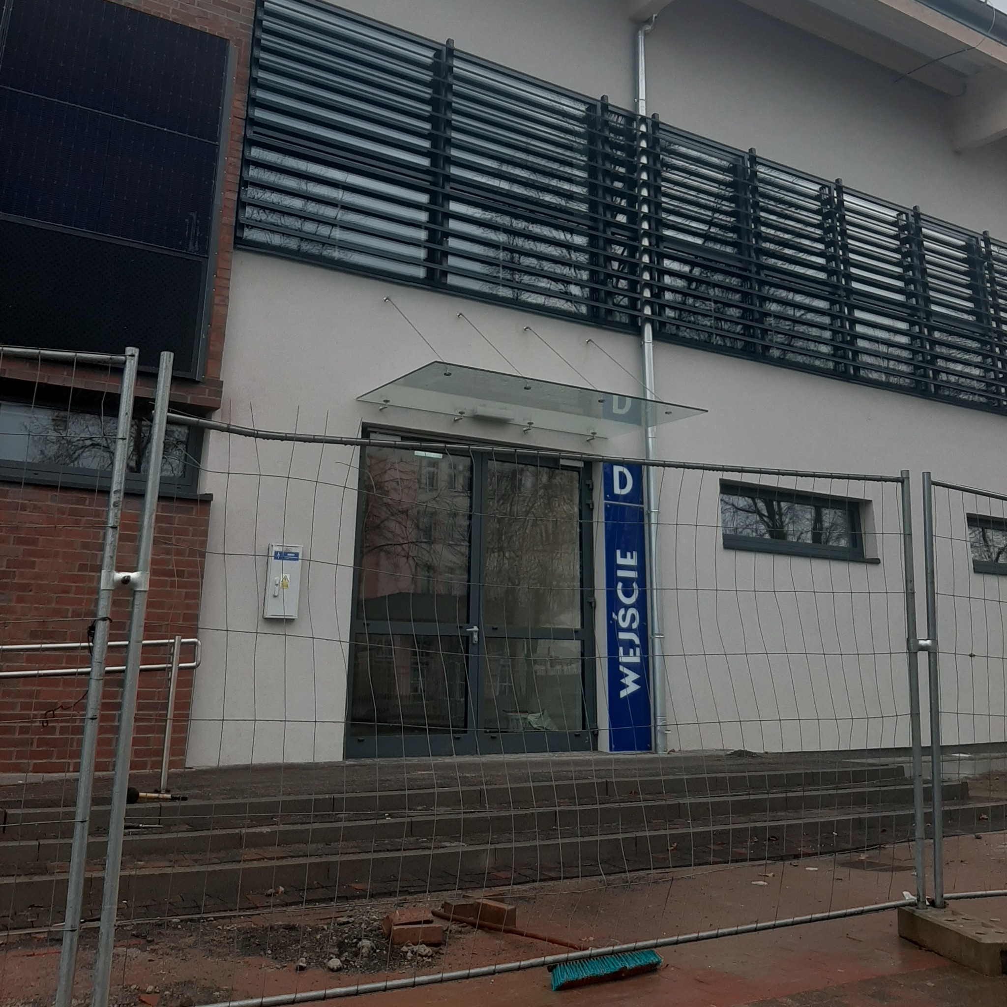 Wejście do budynku z osłoną przeciwsłoneczną nad oknami, zabezpieczone tymczasowym ogrodzeniem budowlanym, widoczna skrzynka elektryczna na ścianie.