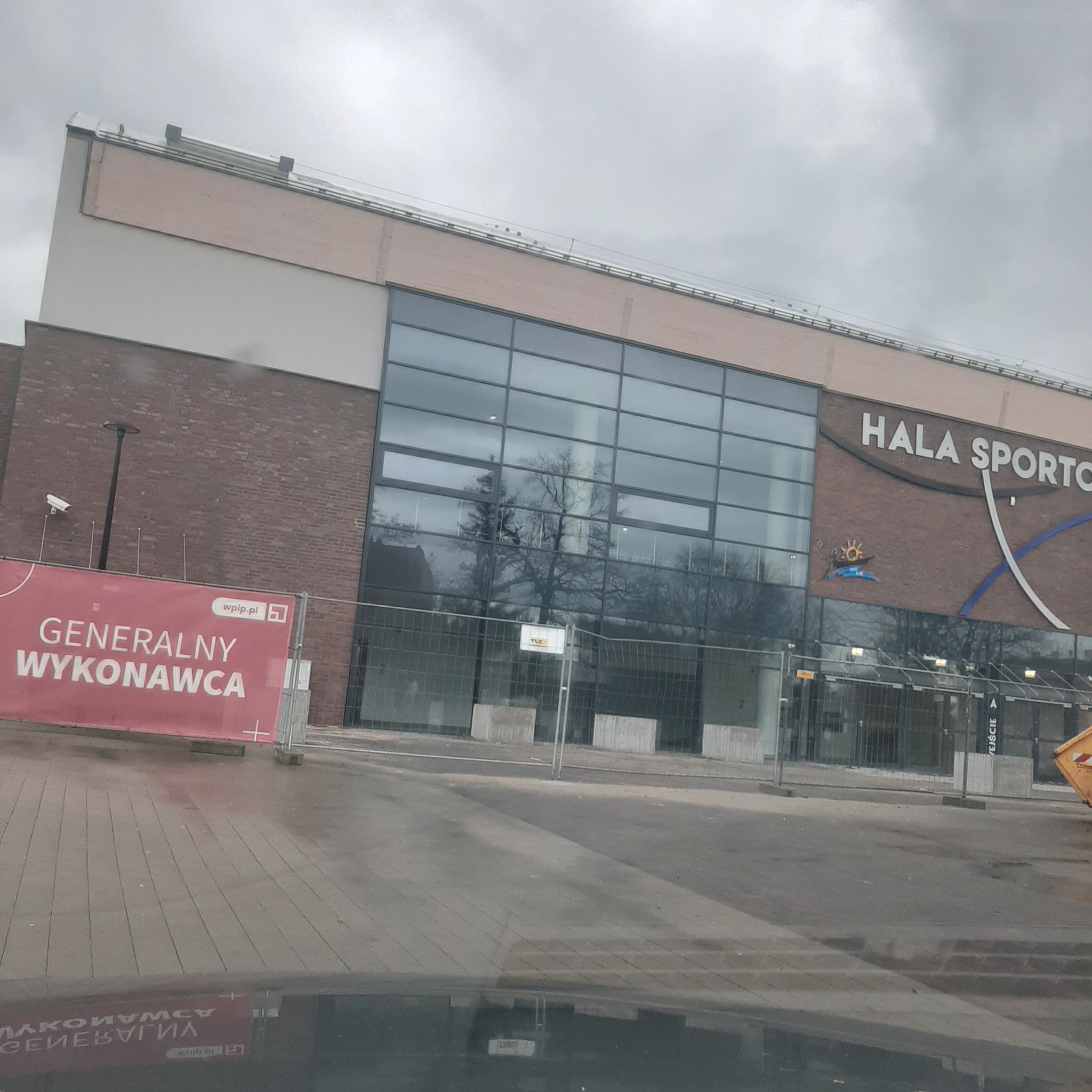 Hala sportowa w trakcie budowy lub remontu, widoczny baner z napisem 'Generalny Wykonawca', szara kostka brukowa na pierwszym planie, metalowe ogrodzenie budowlane.