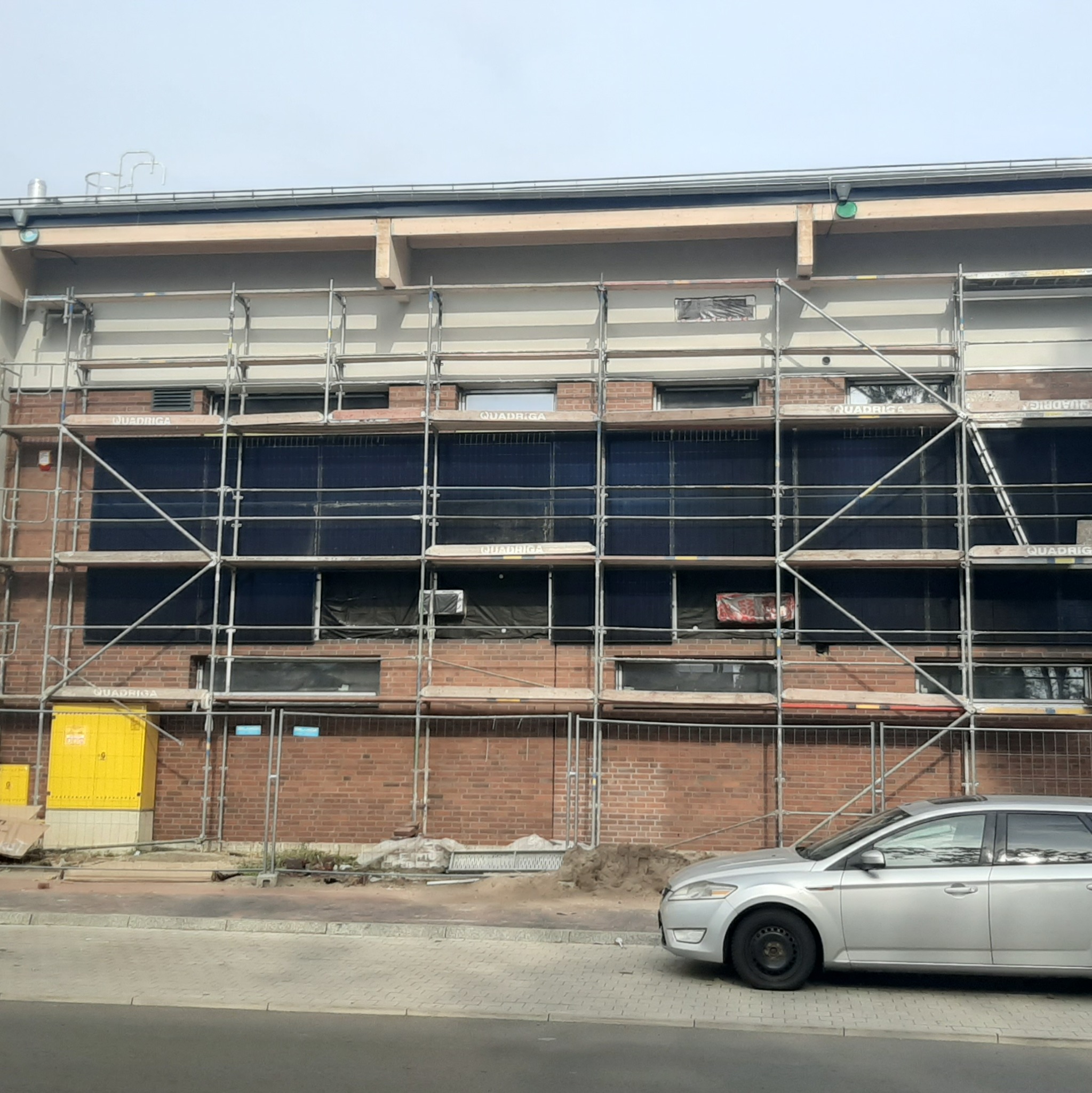 Budynek z cegły w trakcie ocieplania, osłonięty rusztowaniem, widoczne okna zabezpieczone folią, żółta skrzynka elektryczna i zaparkowany srebrny samochód.