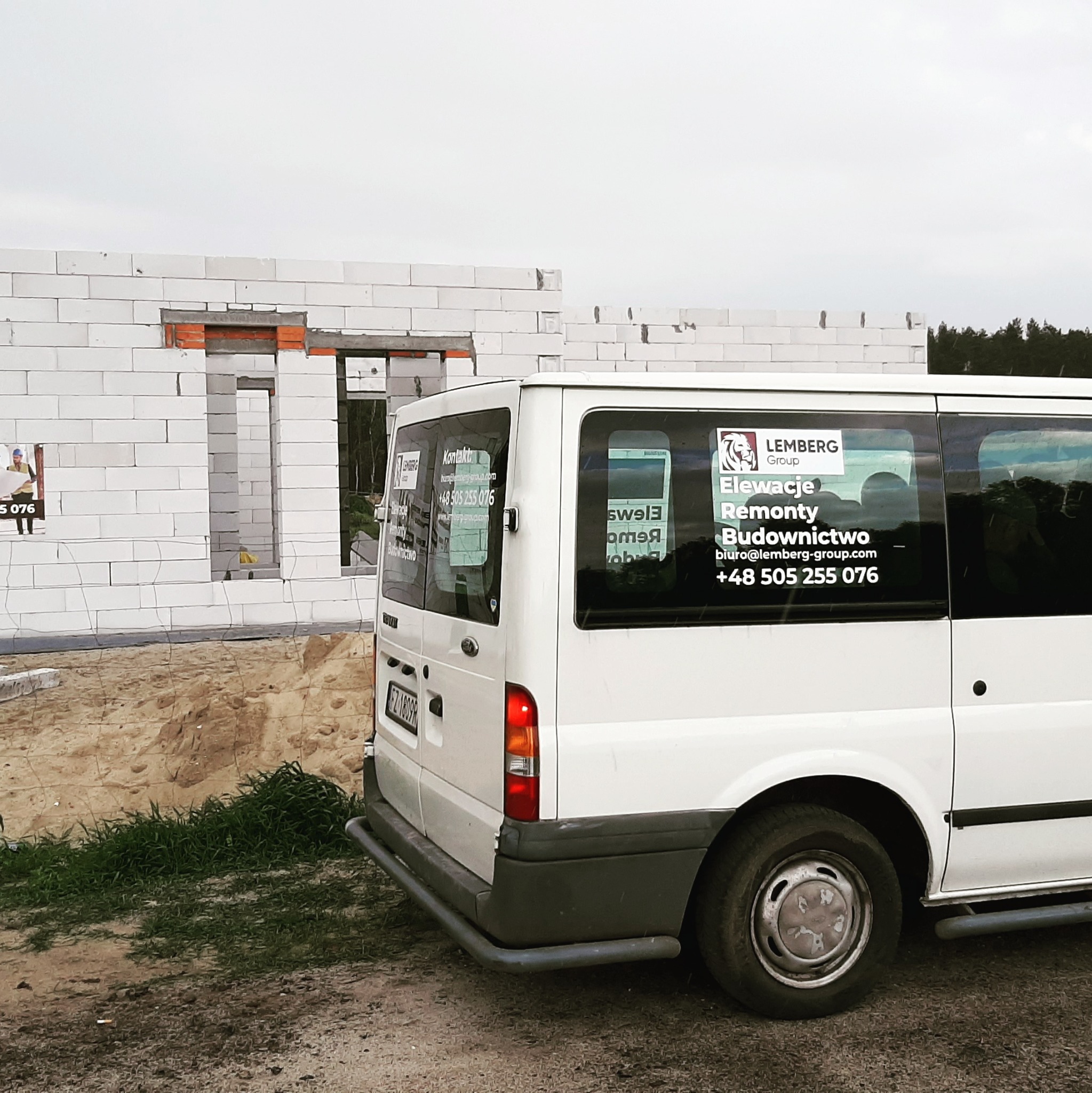 Budynek w trakcie wznoszenia ścian z białych bloczków, na pierwszym planie biały bus z naklejkami reklamowymi firmy budowlanej.