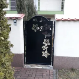 Czarna furtka z wycinanymi motywami kwiatowymi, osadzona między dwoma słupkami ogrodzenia, na tle domu z balkonem.