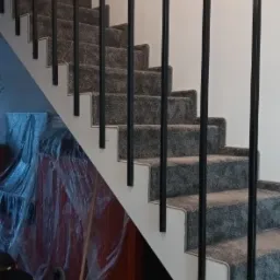 Schody wewnętrzne wyłożone szarą wykładziną z czarną, metalową balustradą, widoczne prace wykończeniowe w tle, w tym folia ochronna i prowizoryczne oświetlenie żarówką.