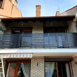 Nowoczesna balustrada balkonowa z poziomych, czarnych profili na tle dwupiętrowego domu z cegły, widoczna drabina sugerująca niedawny montaż.