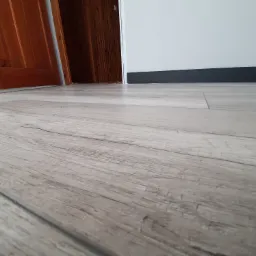 Jasnoszare panele podłogowe z widoczną fakturą drewna, ułożone w kierunku otwartych, drewnianych drzwi, z czarną listwą przypodłogową przy białej ścianie.