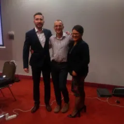 Trzy osoby pozują do zdjęcia na tle ściany podczas konferencji, mężczyzna w garniturze i koszuli bez krawata obejmuje ramieniem mężczyznę w koszuli w paski, obok stoi uśmiechnięta kobieta...